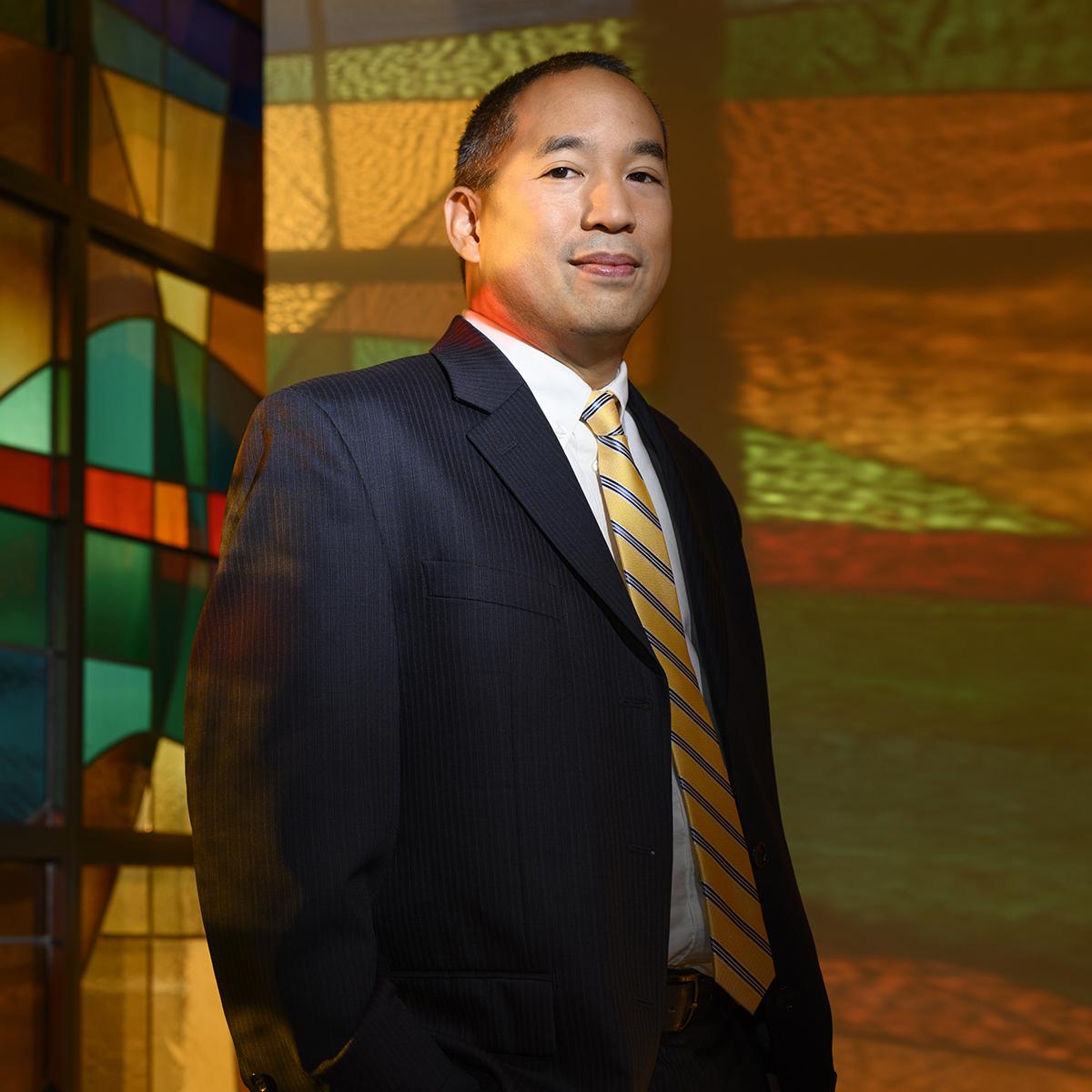 Photo of a smiling man in a suit, st和ing in front of colorful stained glass
